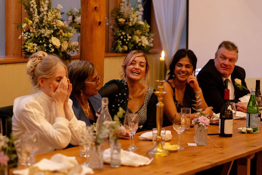 Wedding guests enjoying dinner and conversation at a long wooden table with warm lighting.