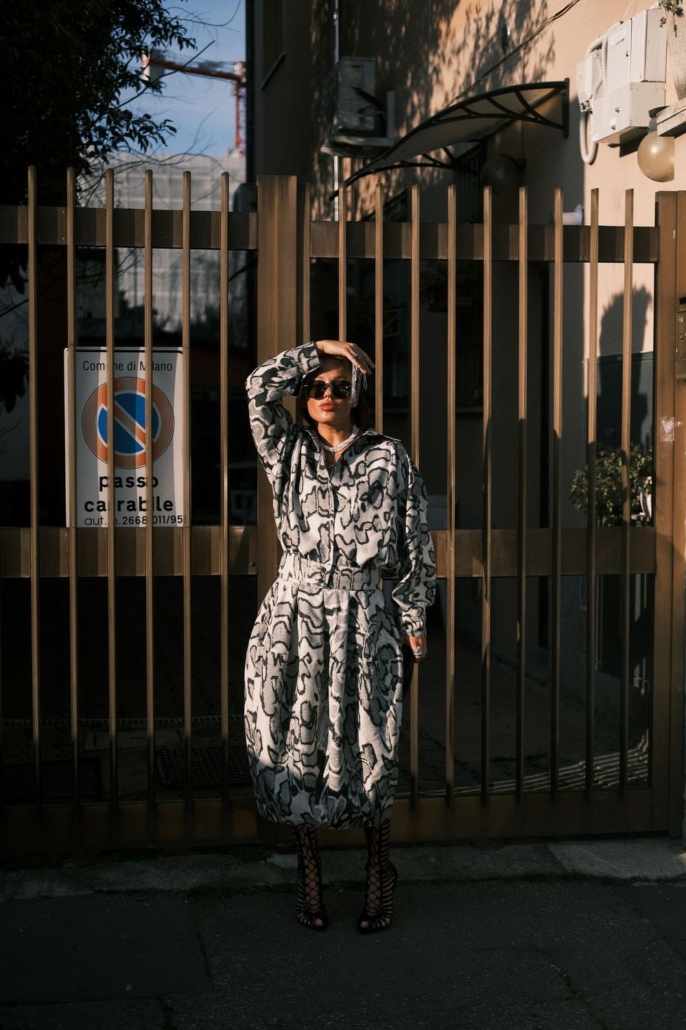 A person in a black and white patterned coat stands against a dark fence at dusk.