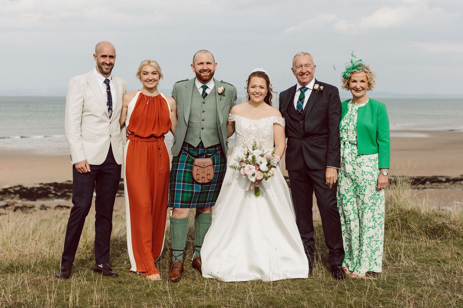 A group of wedding guests pose together on a beach with the ocean in the background, wearing formal attire in vibrant colors.