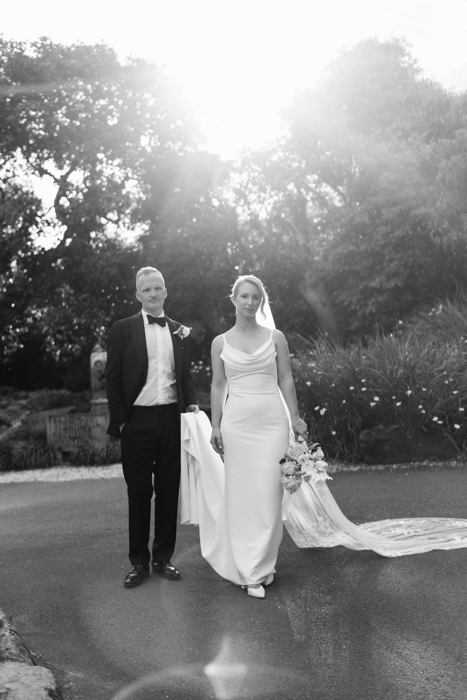 A bride and groom walk together in a garden setting with sunlight streaming through the trees creating a romantic atmosphere.
