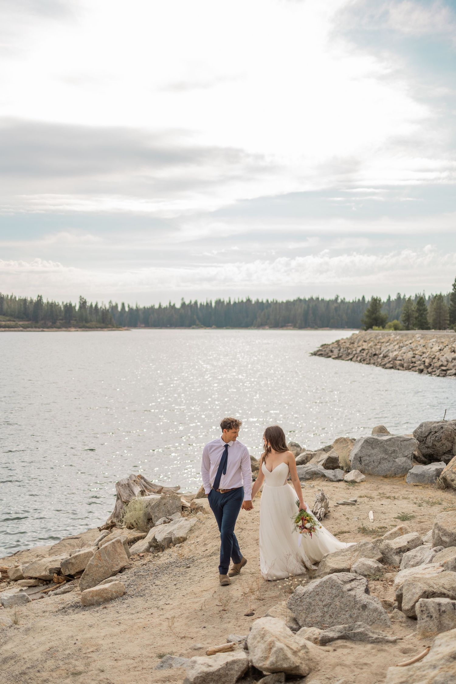 A couple in wedding attire stands on rocky shoreline by a lake with evergreen forest and cloudy sky in background.