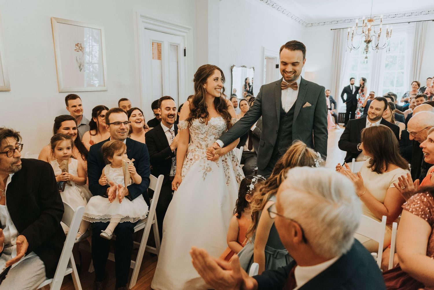 A joyful couple walks down the aisle at an indoor wedding ceremony while guests smile and celebrate their union.