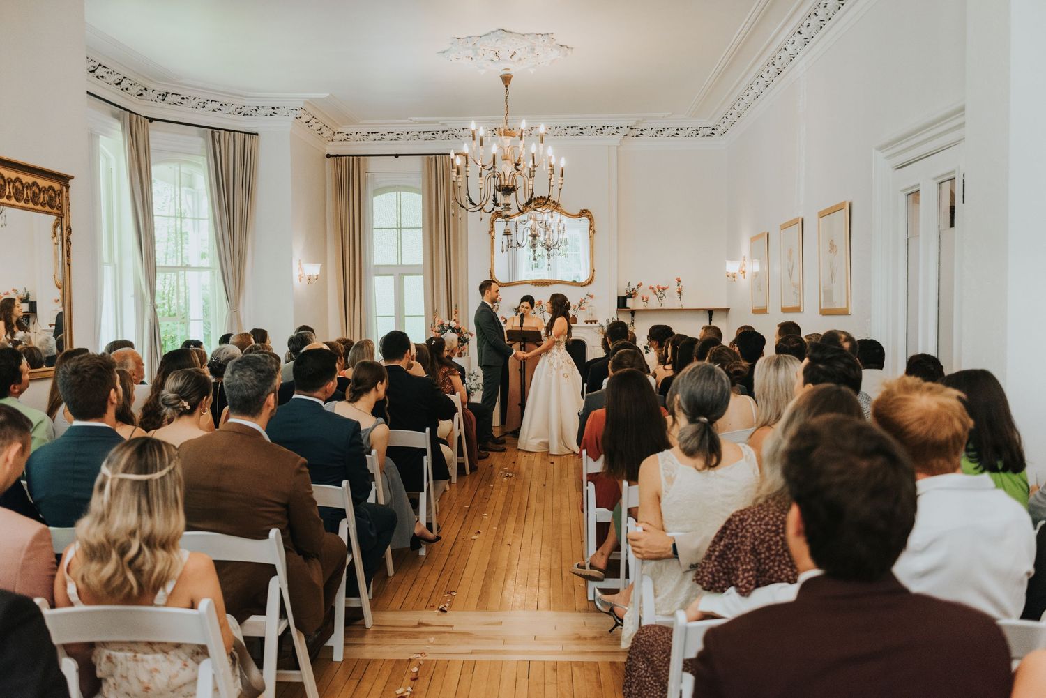 Wedding guests stand in an elegant white ballroom with ornate crown molding and a crystal chandelier during a ceremony.