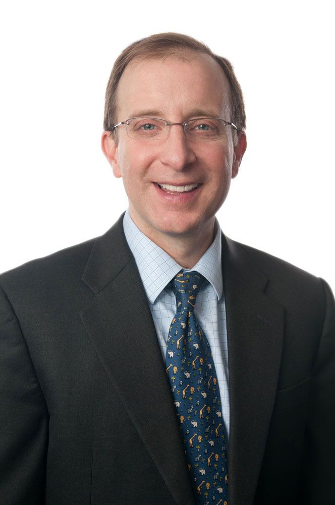 Middle-aged man with glasses wearing dark suit and patterned tie smiling against white background.