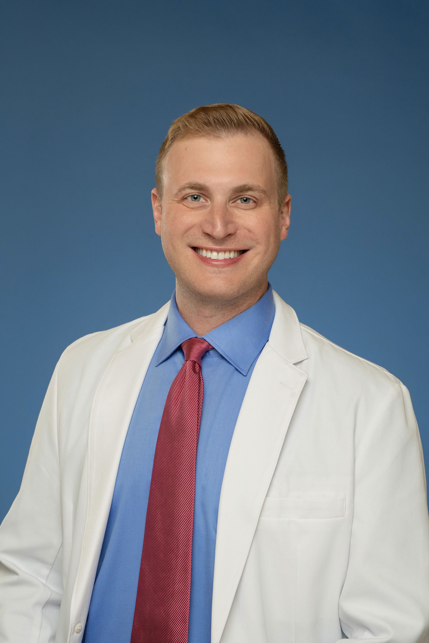 Smiling male doctor in white lab coat and red tie posing against blue background.