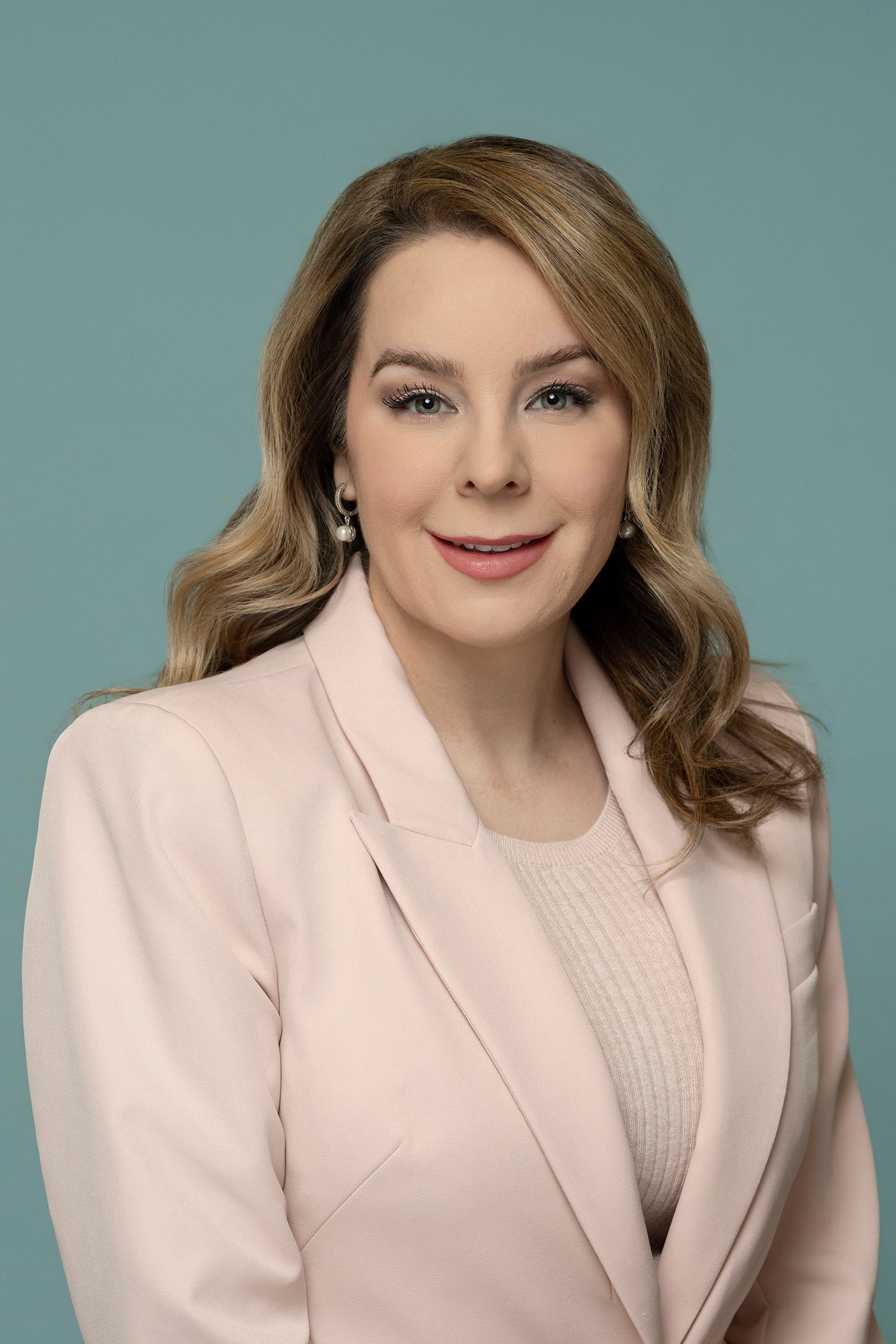 Elegant woman with wavy blonde hair in pink blazer posing for professional portrait on teal background.