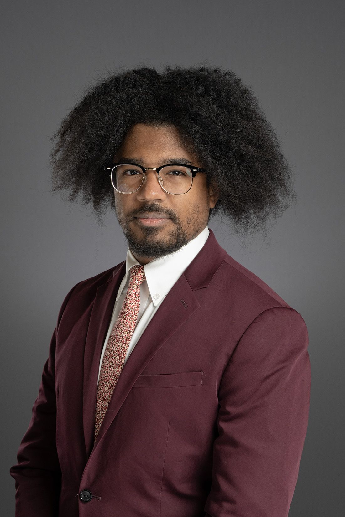 Professional headshot of a man with an afro and glasses wearing a burgundy suit with a floral tie.