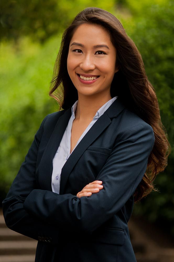 Young Asian woman in navy blazer smiling with arms crossed against green outdoor background.