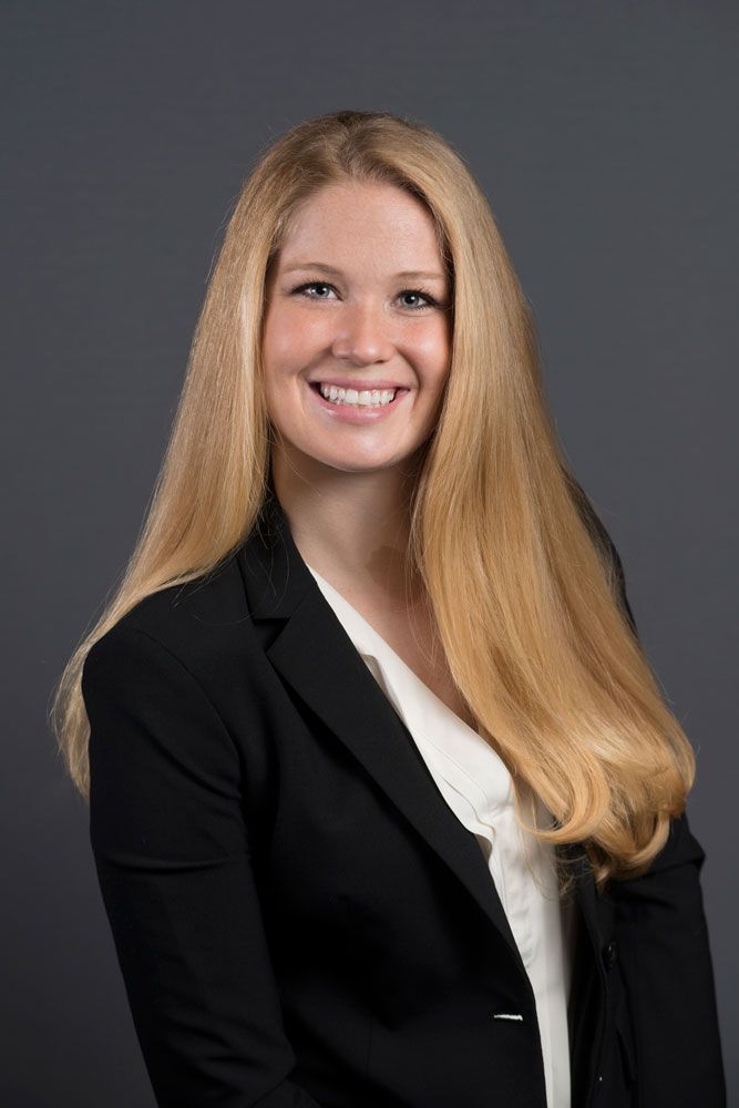 Smiling young professional woman with long blonde hair wearing a black blazer and white blouse against a gray background.