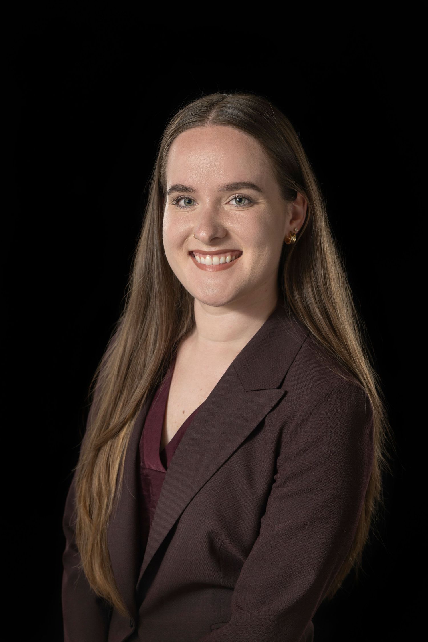 Smiling young woman with long brown hair wearing a maroon blazer against a black background in a professional headshot.