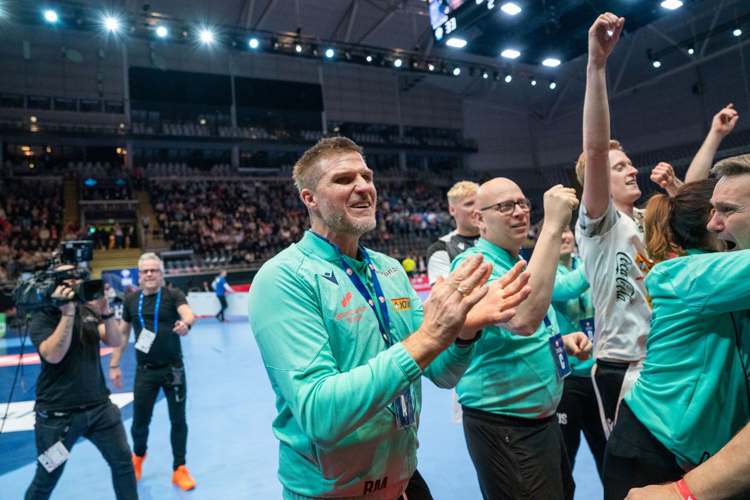 Handball coach celebrates victory with team, clapping and smiling in arena filled with cheering staff in green shirts.