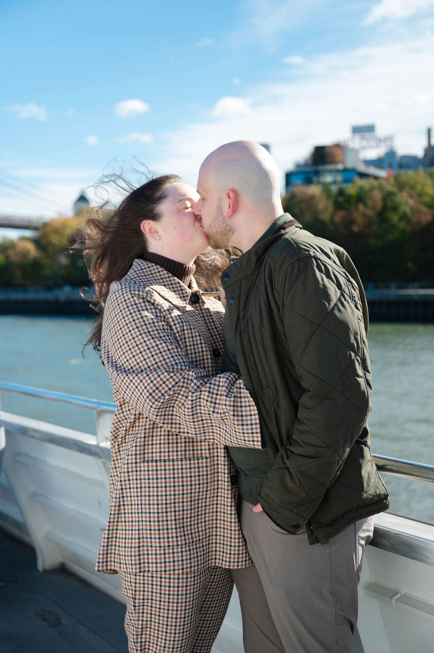 Couple sharing a romantic kiss on a waterfront boardwalk with a bridge and blue sky in the background.