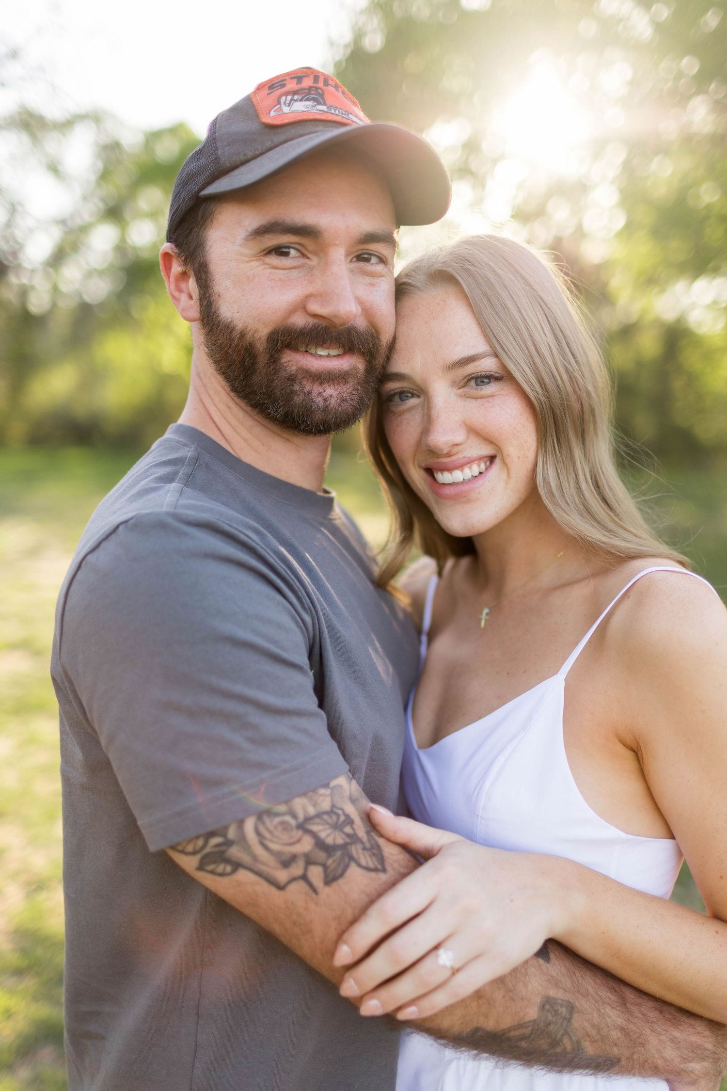 Smiling couple embracing outdoors in golden sunlight, man with beard and cap, woman in white dress with engagement ring in Austin, Texas.