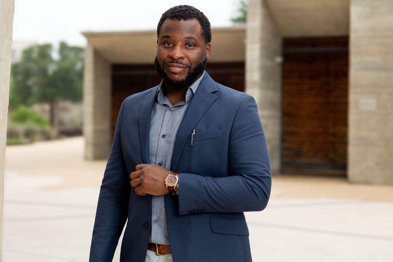 Professional Black man in navy blue blazer and gray shirt poses outdoors near modern building, wearing gold watch.