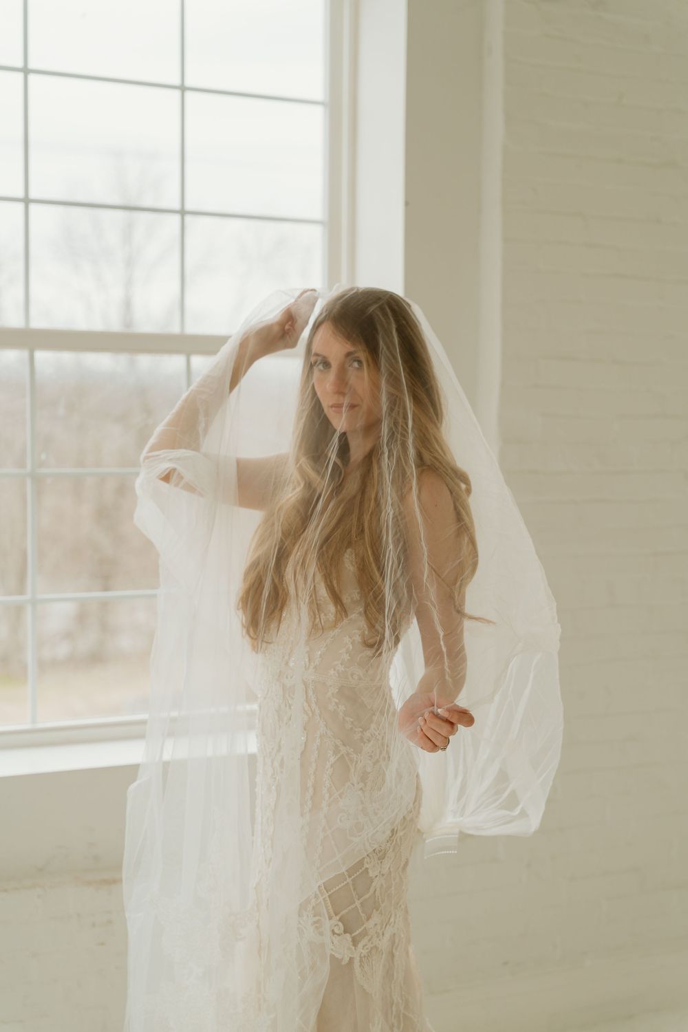 Bride in lace wedding gown lifts flowing veil near large windows in bright white studio space.
