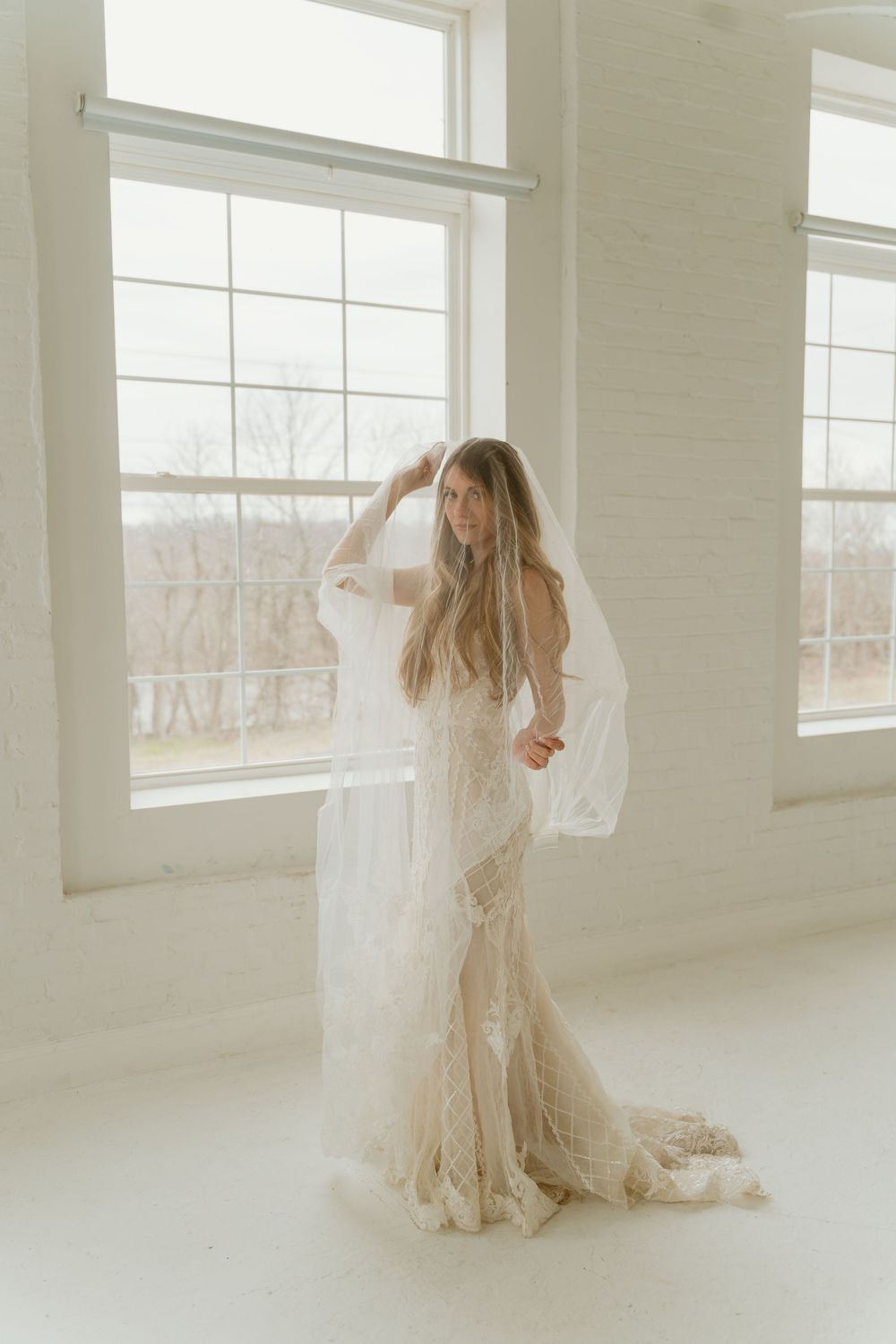 Full-length bridal portrait of woman in fitted lace gown with flowing veil in airy white studio.