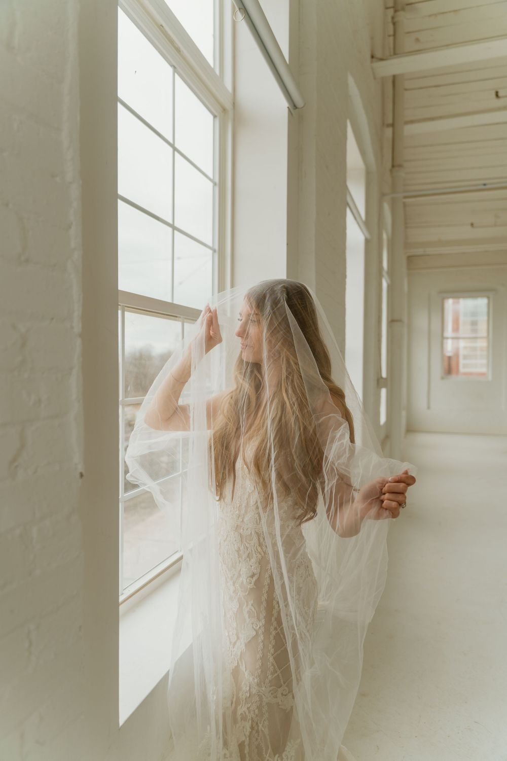 Bride in lace wedding gown and flowing veil gazing out large white-framed windows in a bright airy space.