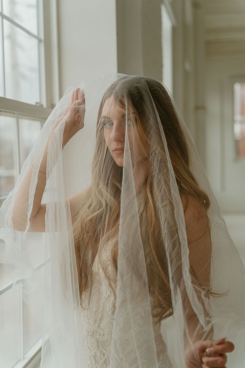 Bride draped in sheer tulle veil covering her face, wearing lace wedding dress near bright windows.