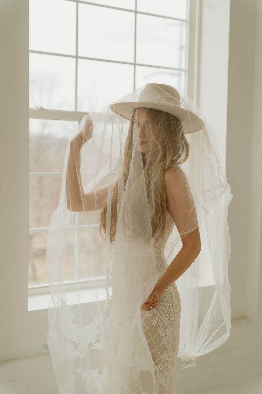 Boho bride in lace gown and wide-brim hat with flowing veil standing by large window in white room.