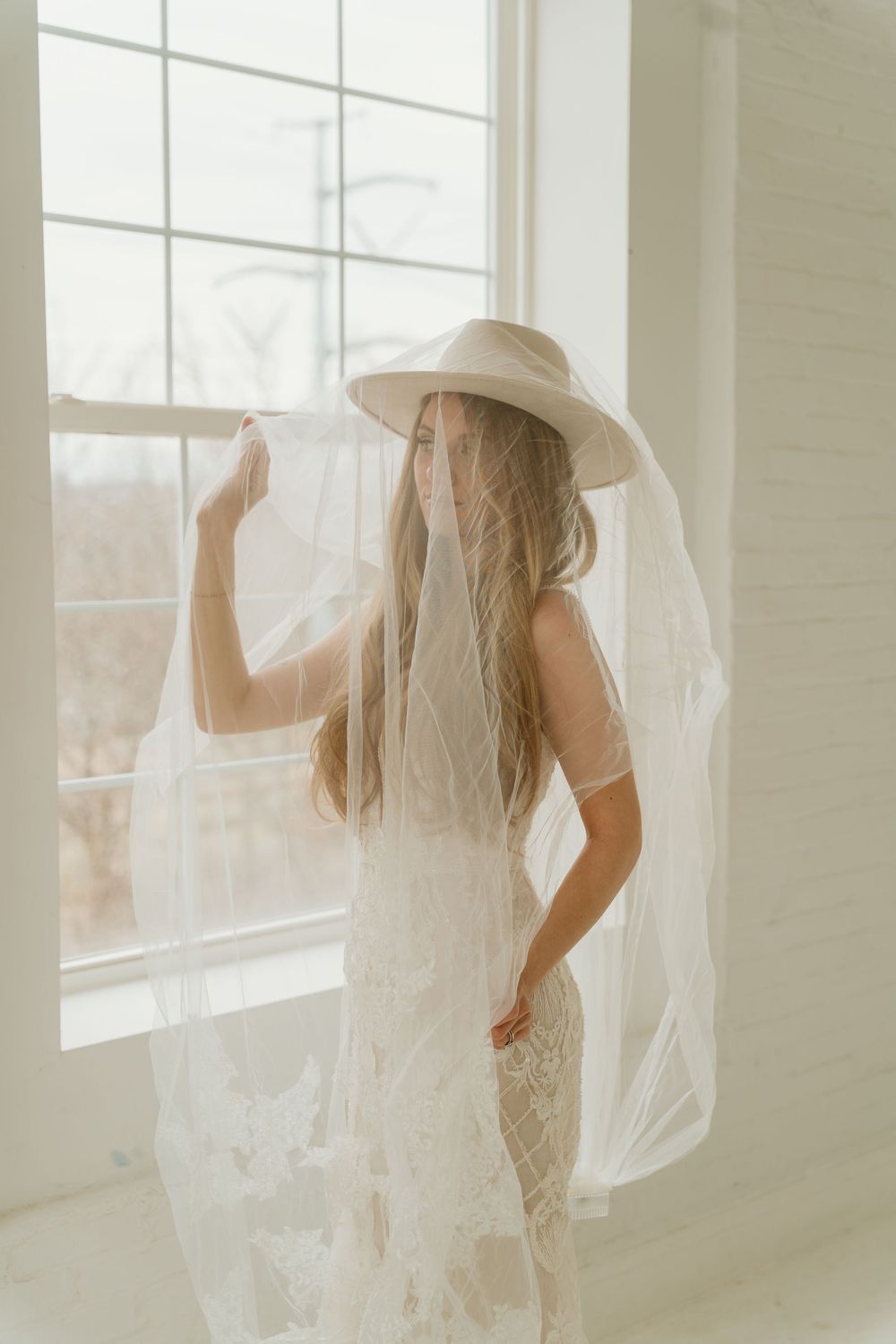 Back view of bride in lace wedding dress and wide-brim hat with sheer veil by bright window.