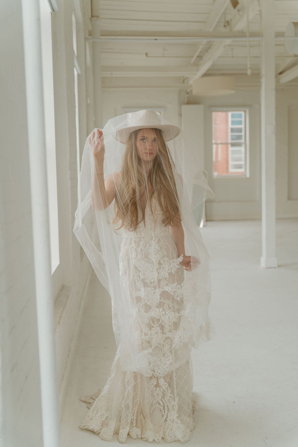 Boho bride in lace mermaid gown and wide-brim hat holding flowing veil in bright white venue space.