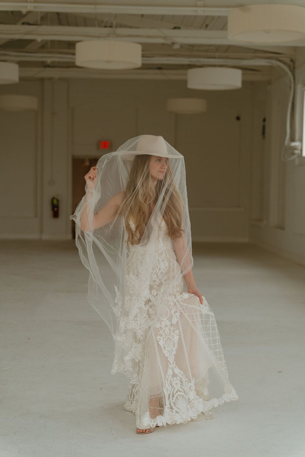 Bride in lace wedding dress and wide-brim hat twirling with flowing veil in bright open venue room.