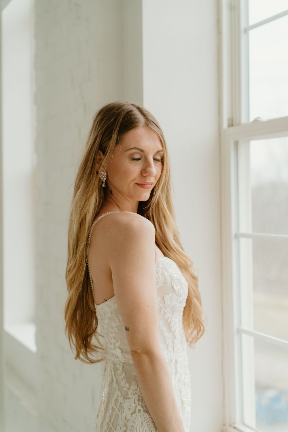 Bride in strapless lace wedding gown with long blonde hair smiling softly beside a sunlit window.