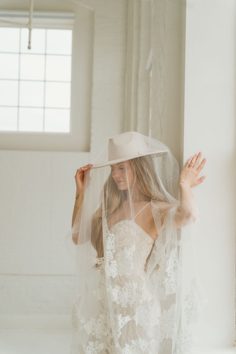 Bride in lace wedding gown and wide-brim hat with sheer veil posing elegantly in a bright white room.