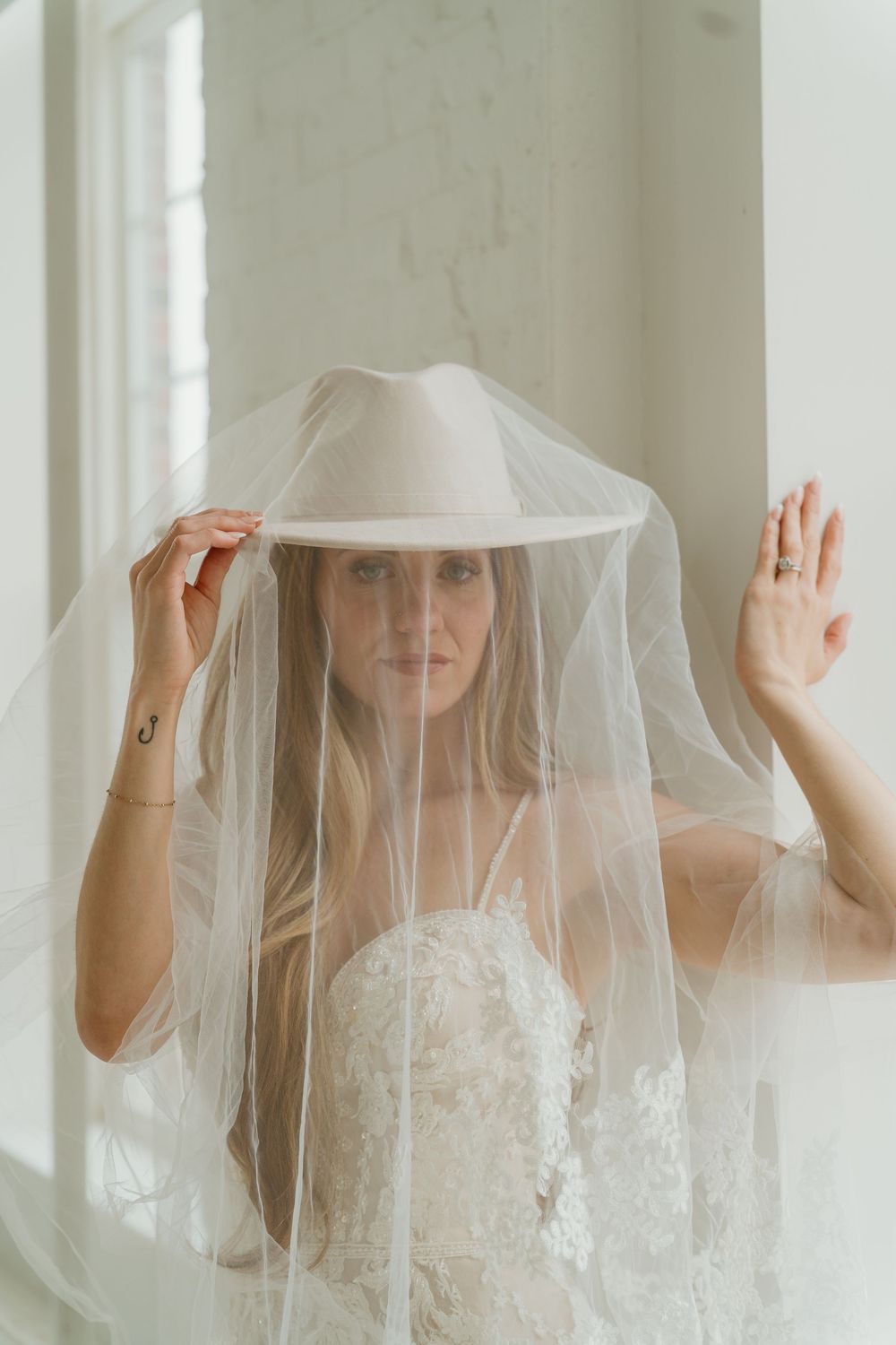 Bride wearing a lace wedding dress with a cowboy hat draped under a sheer veil indoors.