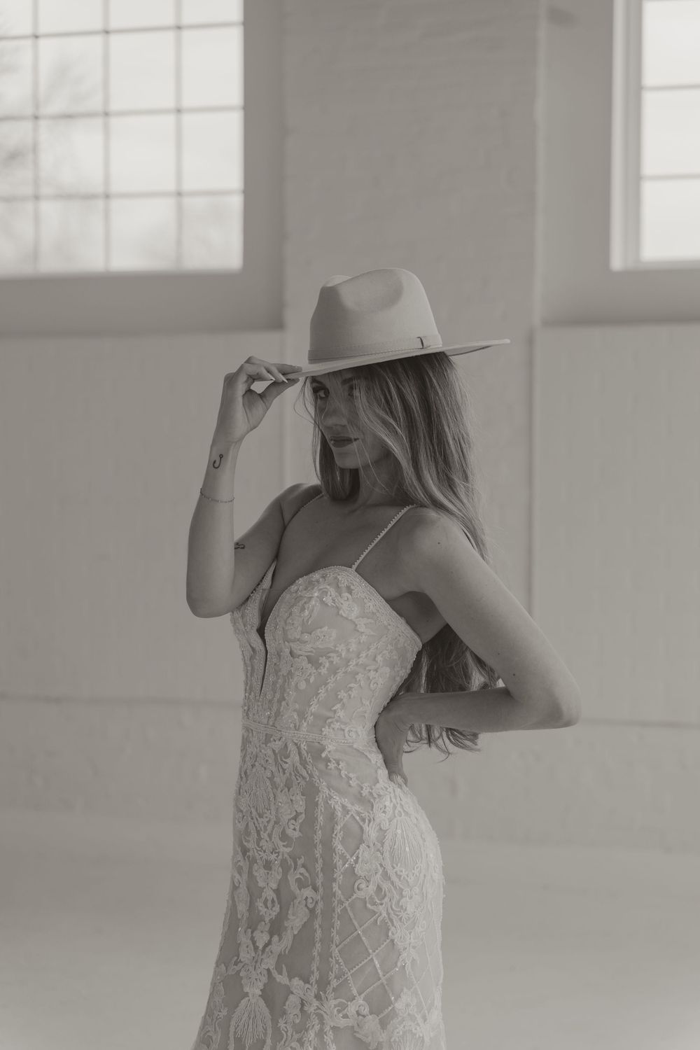 Black and white photo of a bride in a lace gown tipping a wide-brim cowboy hat in an open space.