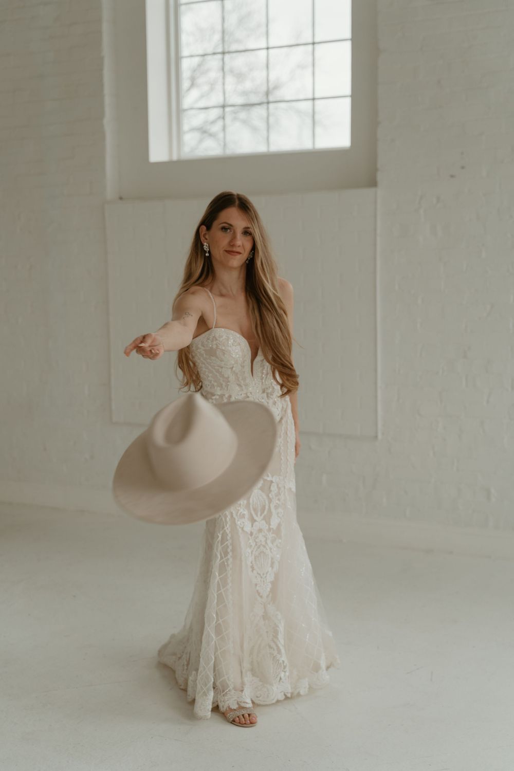 Bride in a strapless lace wedding gown holding a wide-brim hat in a bright white studio.