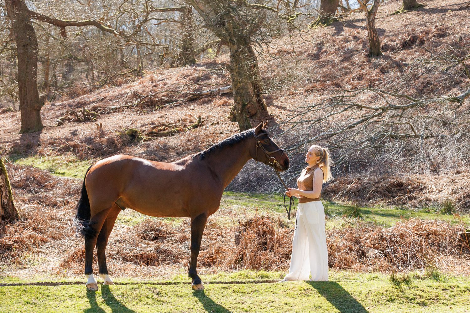 Bride in white gown standing beside a brown horse in a sunlit woodland setting.