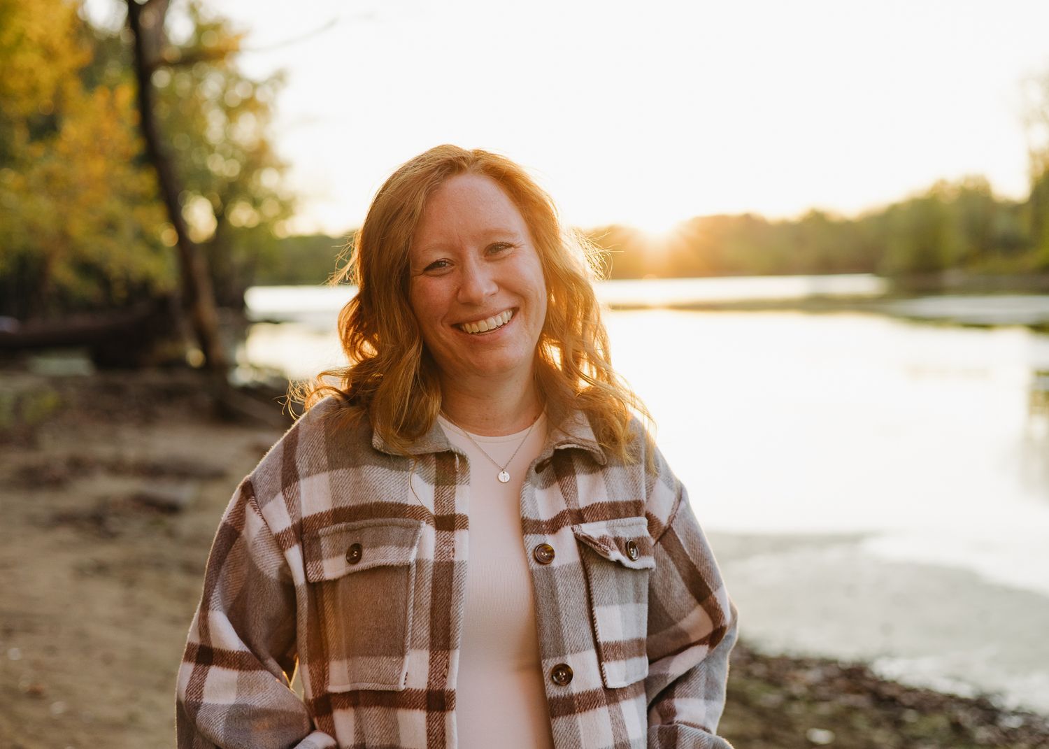 Smiling young woman in plaid jacket standing by a sunlit lake during golden hour.