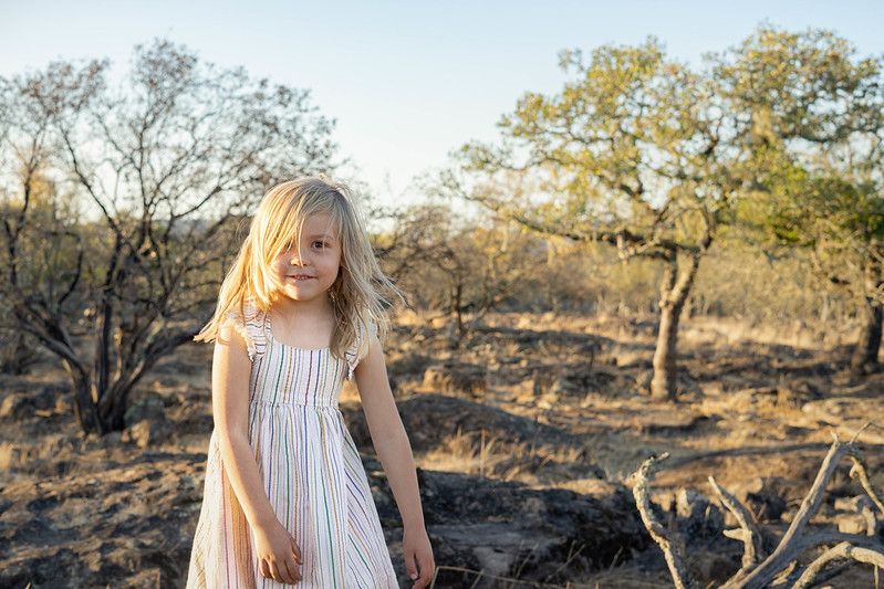 A young child in a white dress stands in a dry, sunlit landscape with scattered trees in the background.