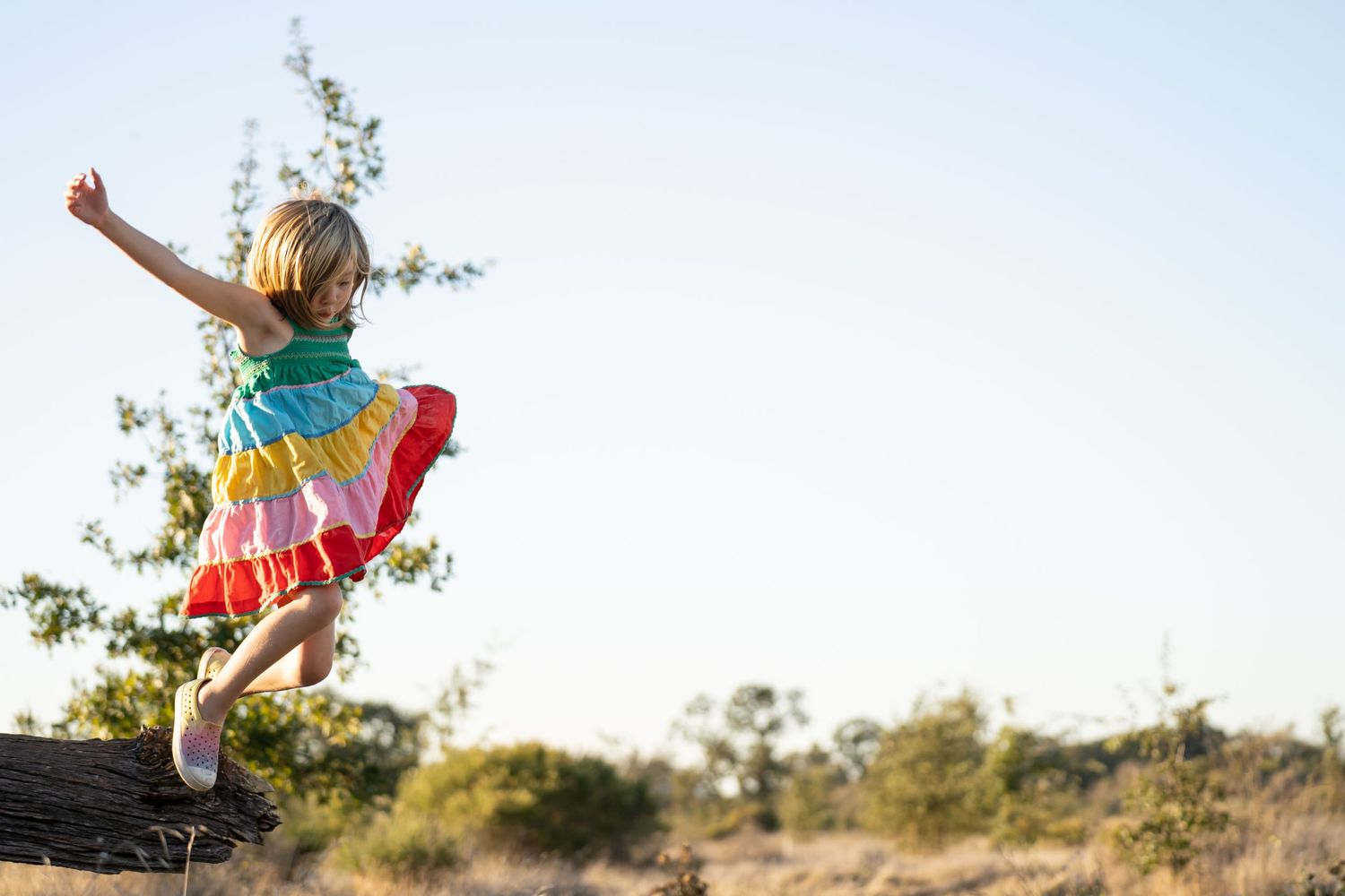 A person in a colorful rainbow dress leaps joyfully from a rock against a sunny outdoor background.