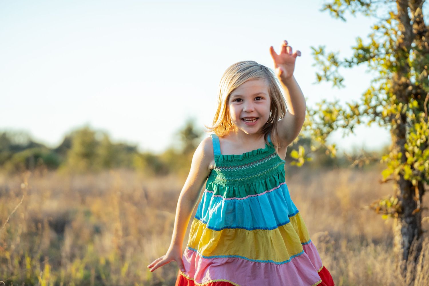 A child wears a colorful tiered rainbow dress while playing joyfully in a sunlit meadow during golden hour.
