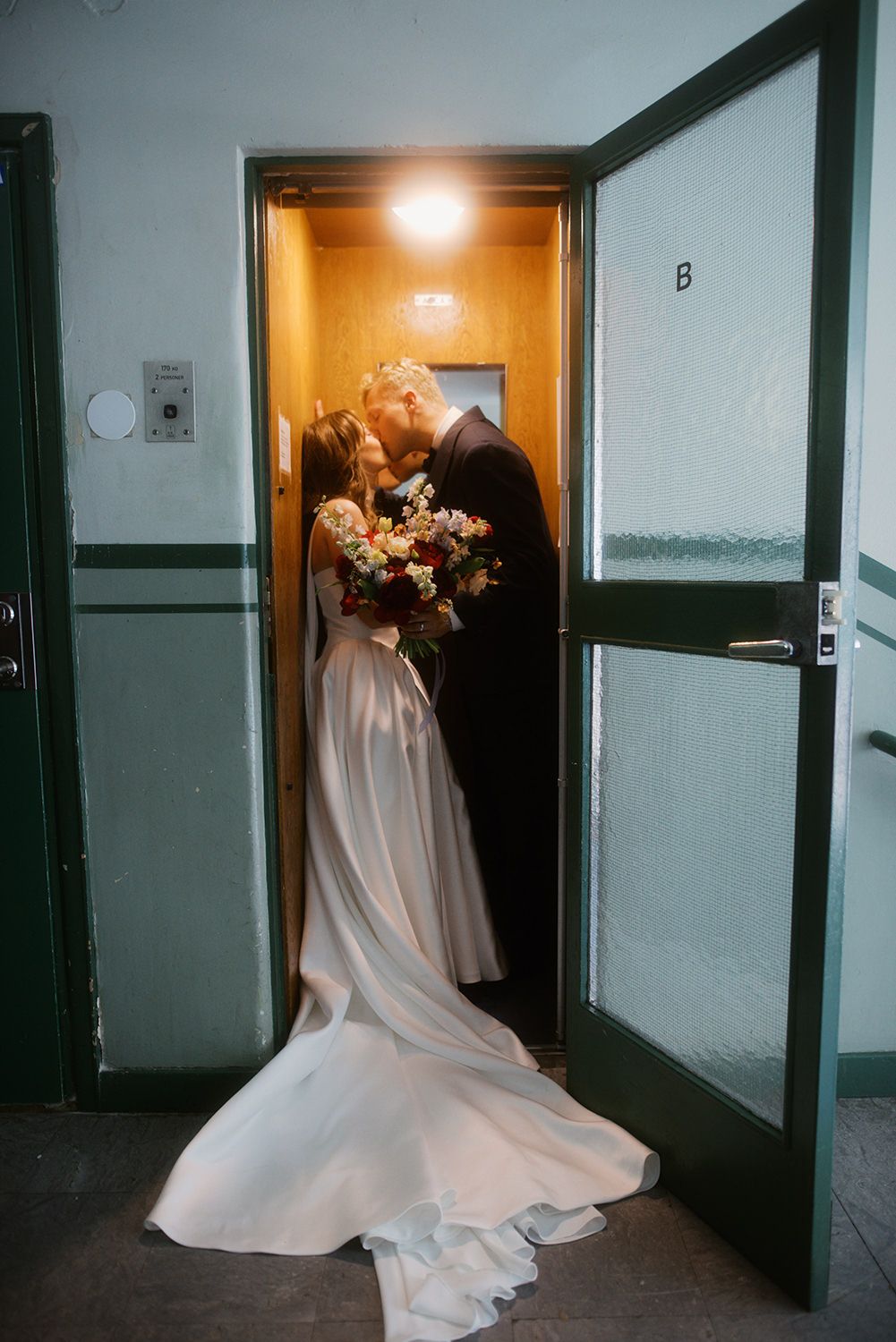 Bride and groom sharing a kiss in a warmly lit elevator doorway, bride holding a colorful bouquet in a flowing white gown.