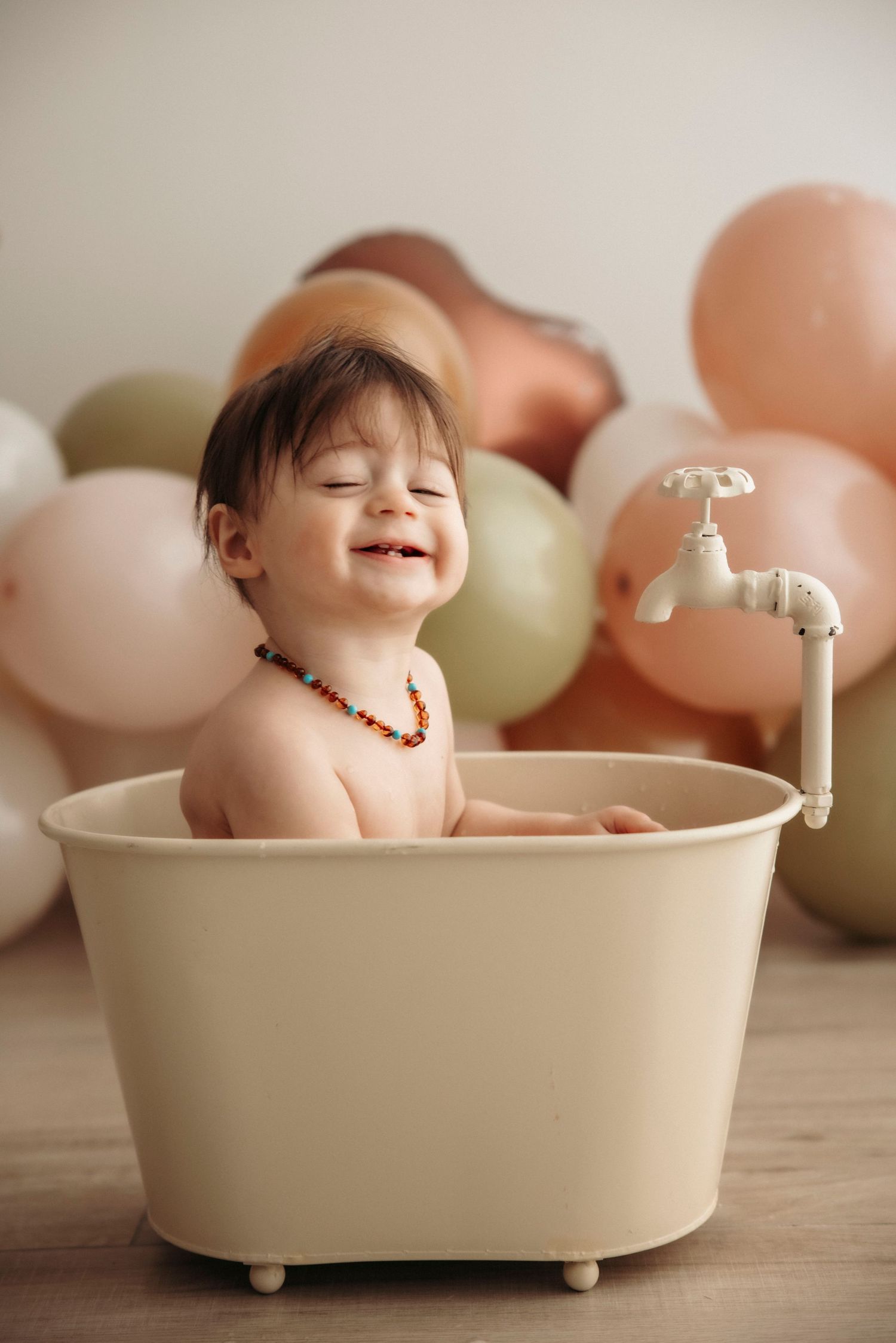 Smiling baby sitting in a small cream bathtub surrounded by pastel balloons, wearing an amber teething necklace.