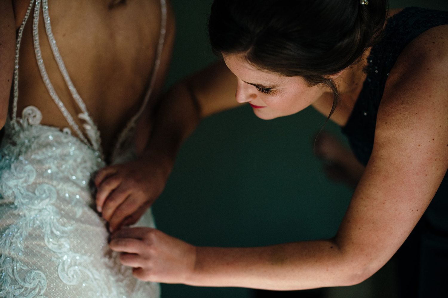 Bridesmaid carefully fastening the back of a beaded lace wedding dress on the bride before the ceremony.