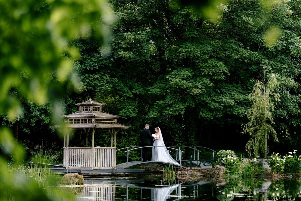 A scenic white gazebo and bridge over a tranquil pond surrounded by lush green trees in a romantic garden setting.
