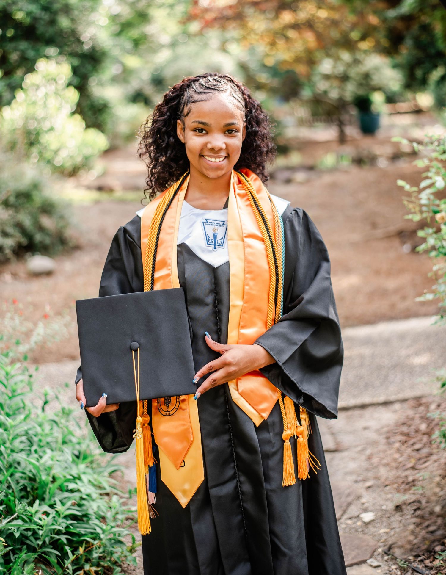 Young woman in black and gold graduation cap and gown smiling outdoors, holding her cap with honor stoles.