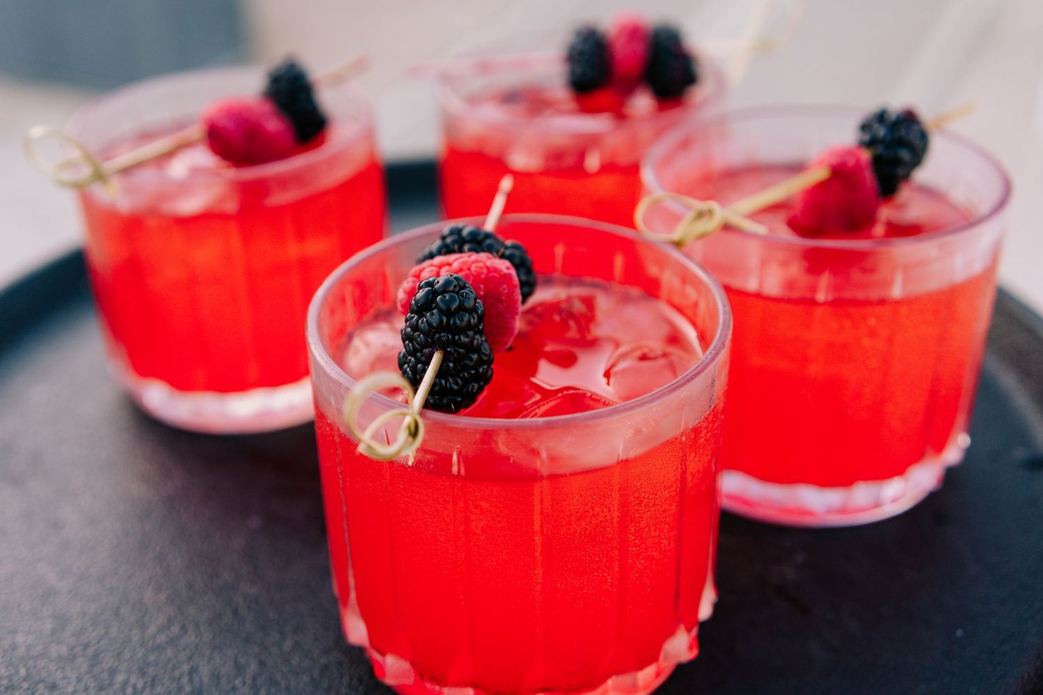Four red cocktails garnished with fresh blackberries and raspberries on skewers, served in rocks glasses on a dark tray.