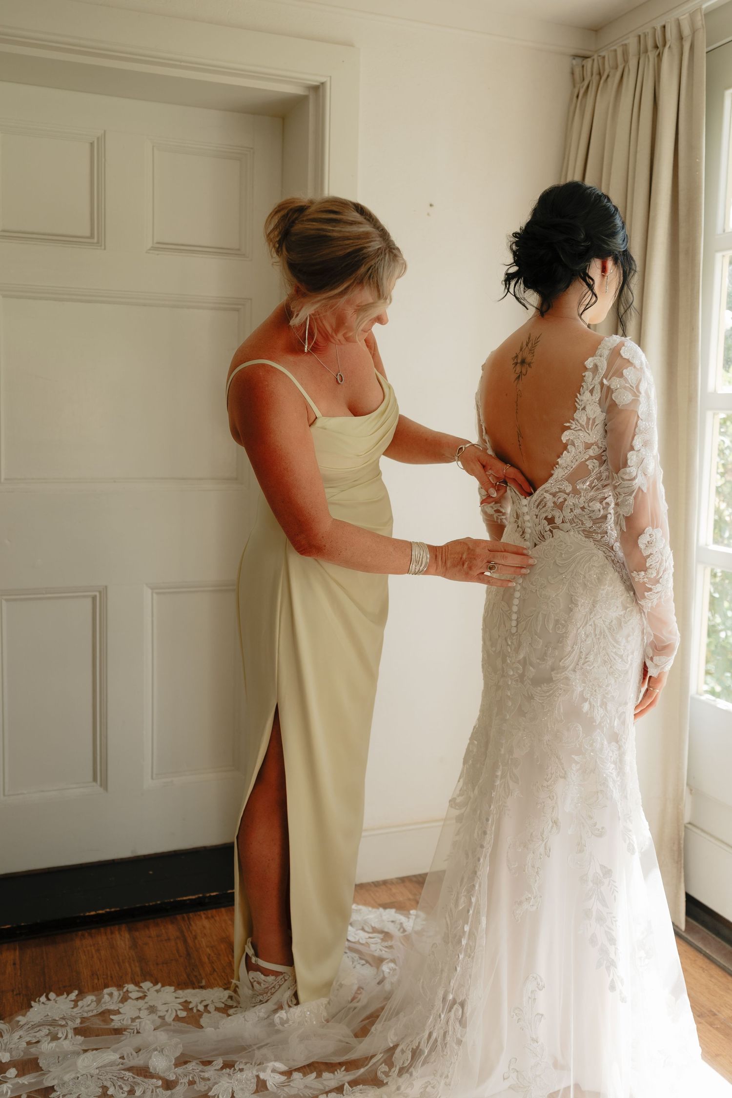 Bridesmaid in yellow dress helping bride in lace wedding gown button up near a window.