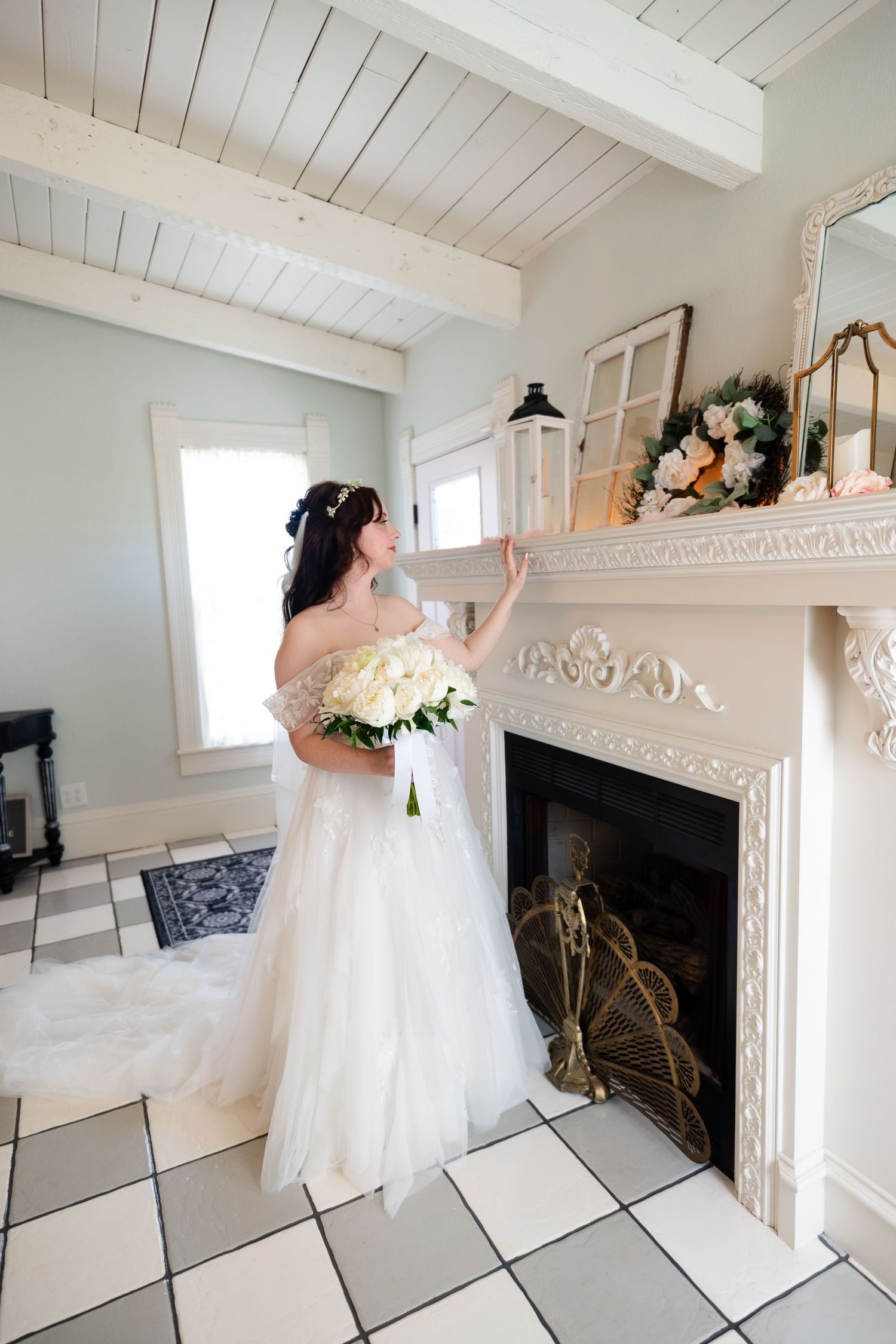 Bride in white off-shoulder gown holding bouquet beside ornate white fireplace in elegant room with checkered floors.