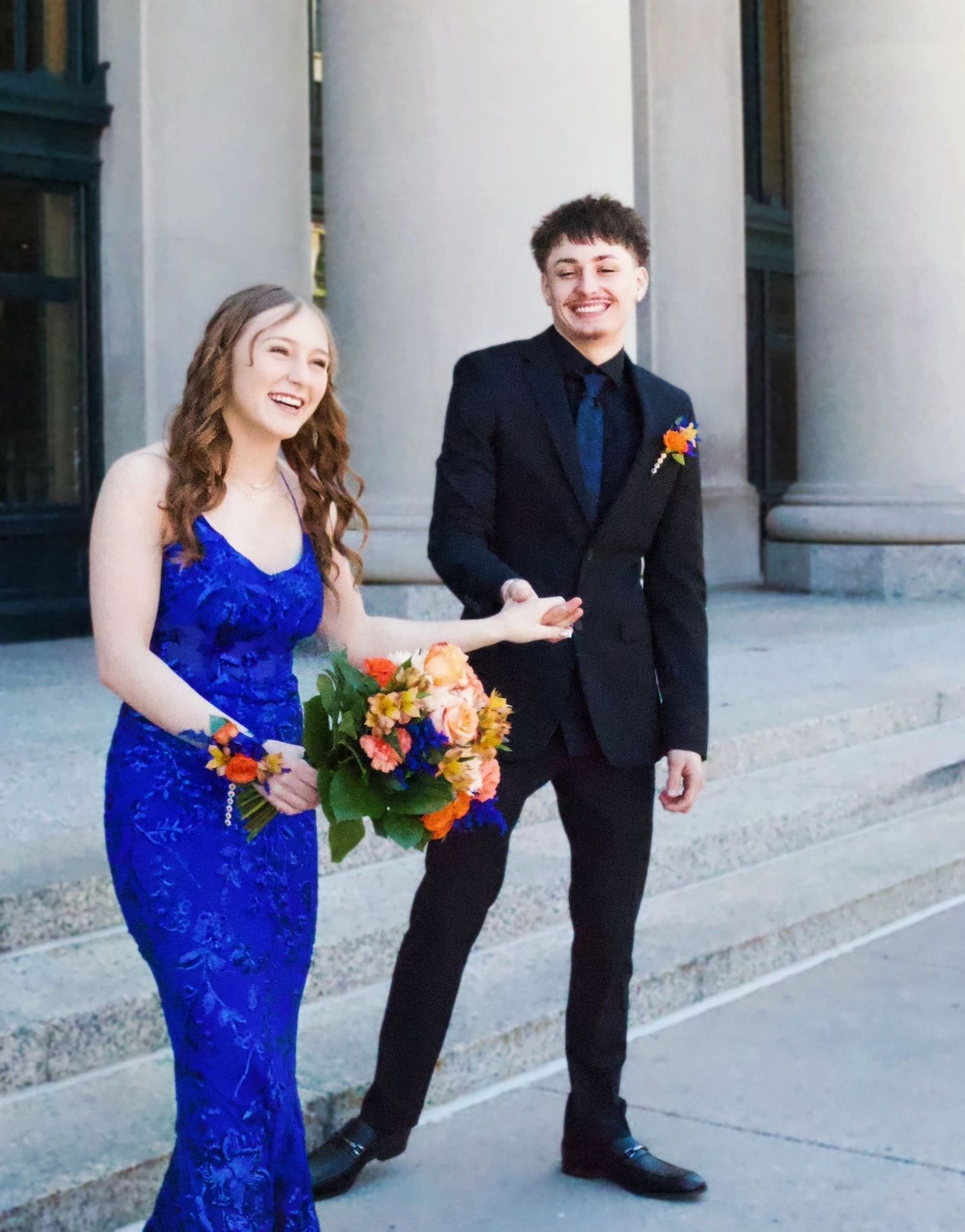 A young couple in prom attire, girl in blue gown holding flowers, boy in black tuxedo with boutonniere, posing outdoors.
