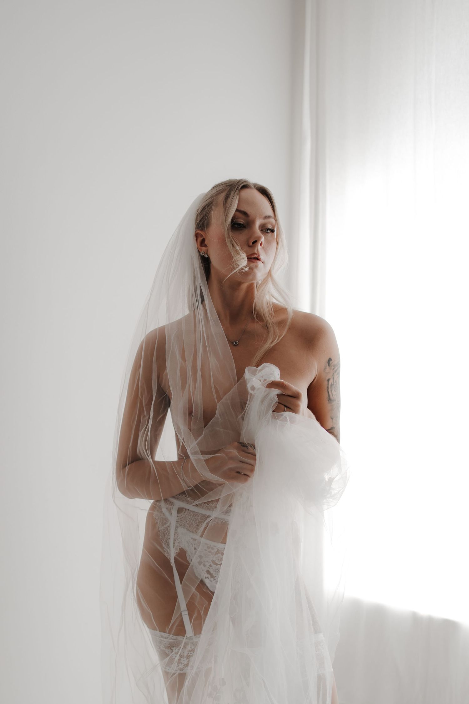 Woman in white lace bodysuit and heels posing against a wall near sheer curtains in a bright white room.