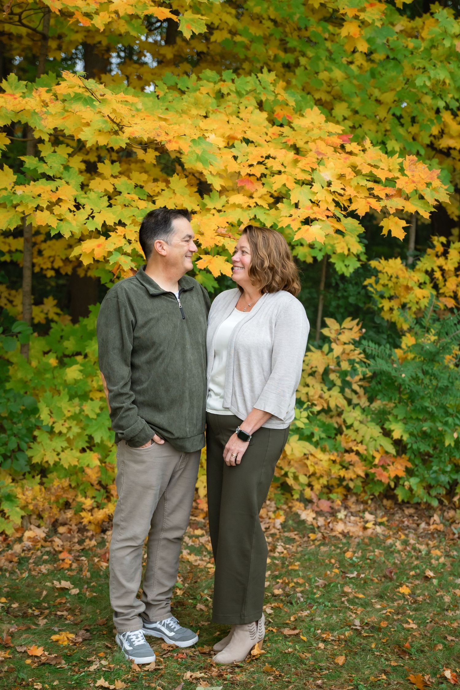 Couple smiling at each other outdoors surrounded by vibrant yellow and green autumn foliage in a park setting.