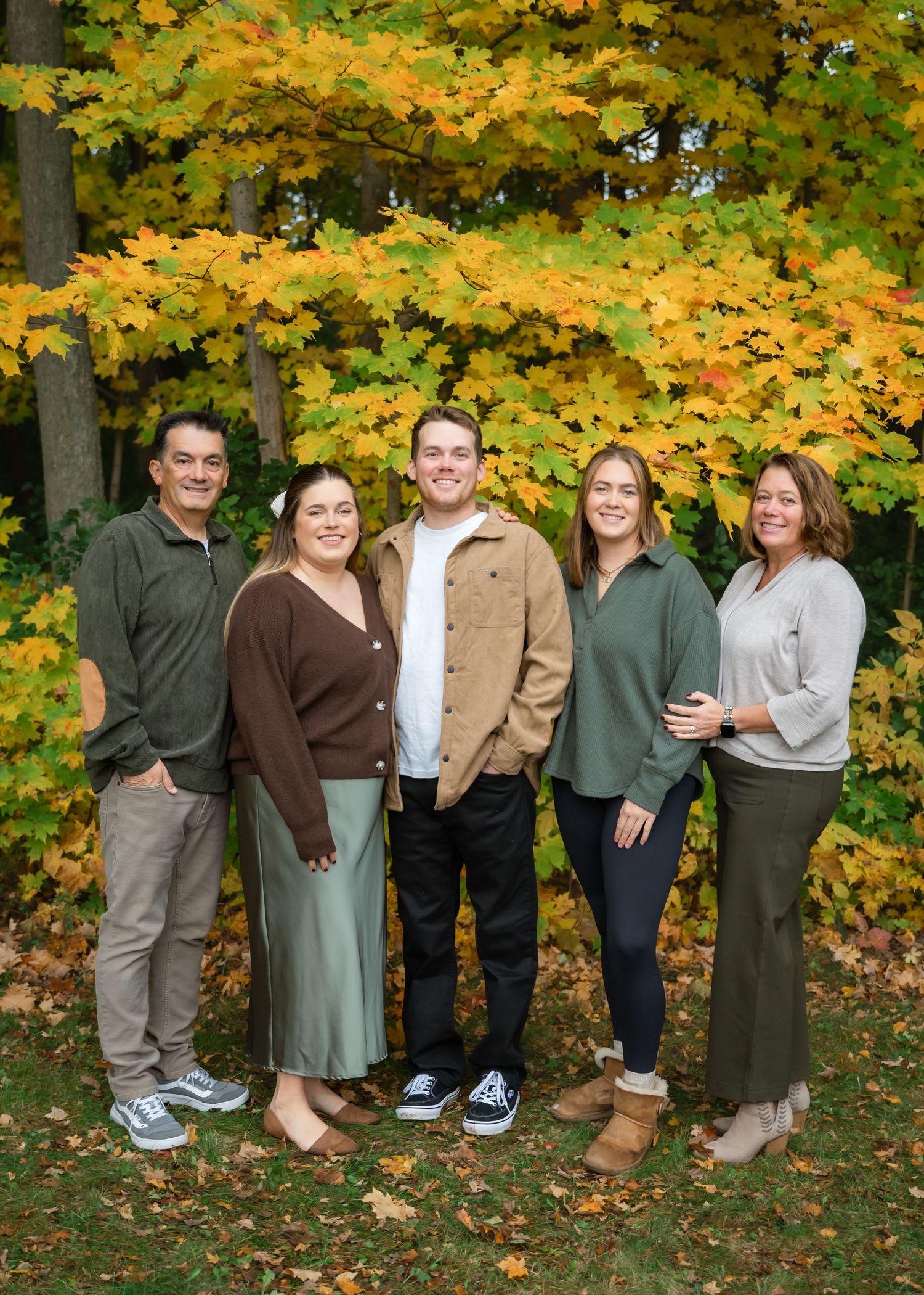 Family of five smiling together outdoors in front of vibrant yellow and orange autumn foliage trees.