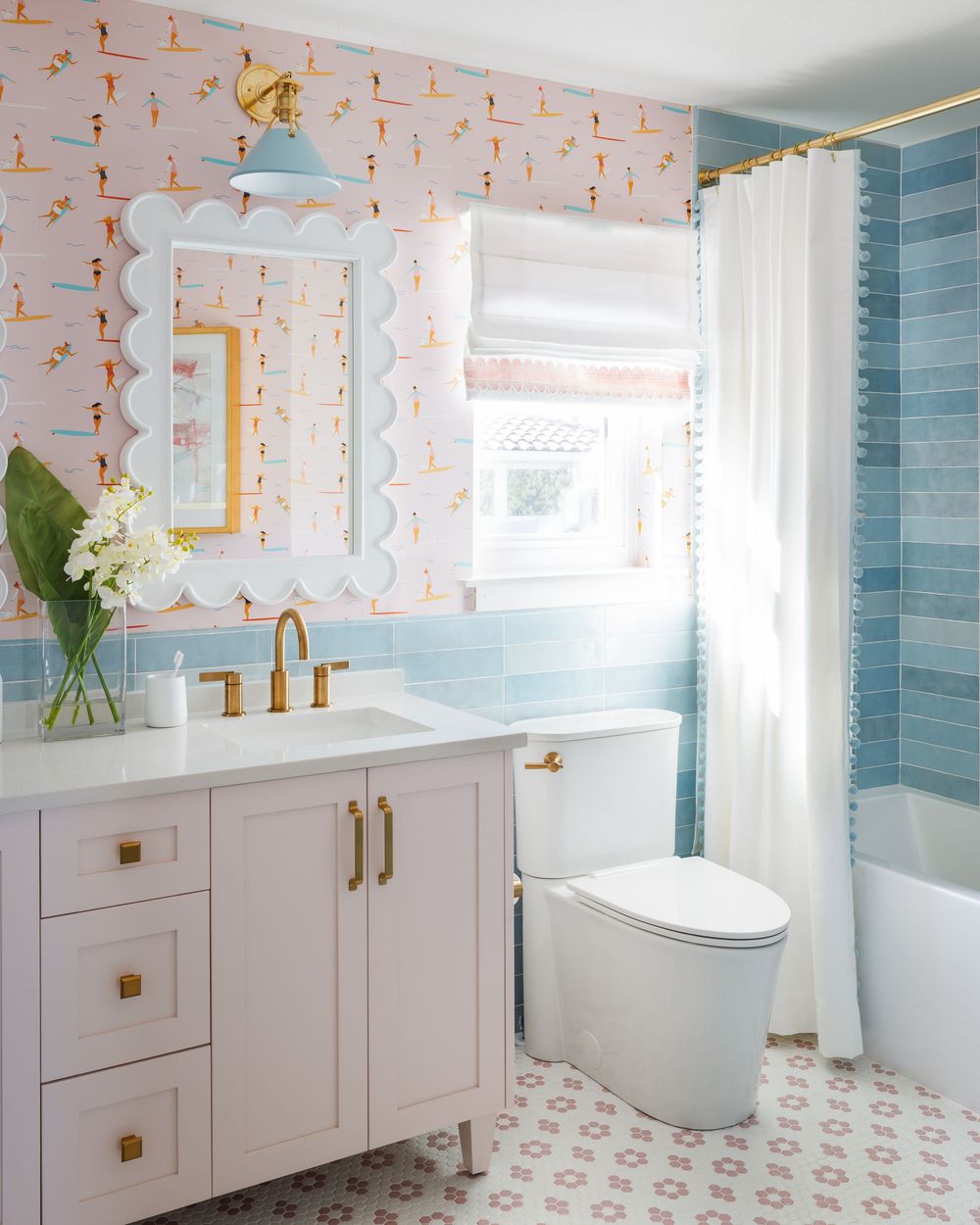 Playful bathroom with pink patterned wallpaper, blue tile tub surround, scallop mirror, and gold fixtures.
