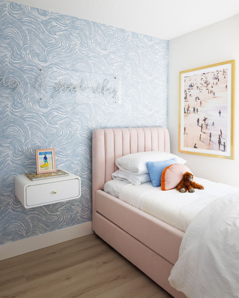 Pink upholstered twin bed with blue wave wallpaper accent wall and beach photography art in a child's bedroom.